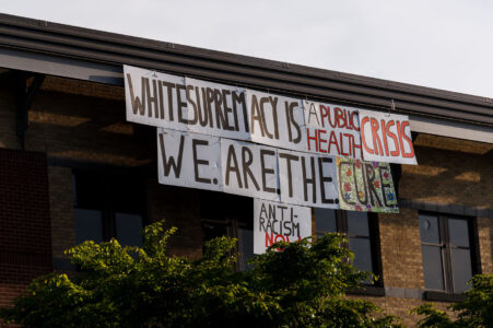 A sign reading "White supremacy is a public health crisis. We.Are.The.Cure. Anti-Racisim NOW" hanging over the side of the Minneapolis Police Third Precinct on May 31, 2020.