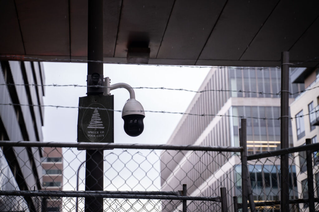 A sign reading "Spruce Up Your City" next to a surveillance camera behind barbed wire in downtown Minneapolis.