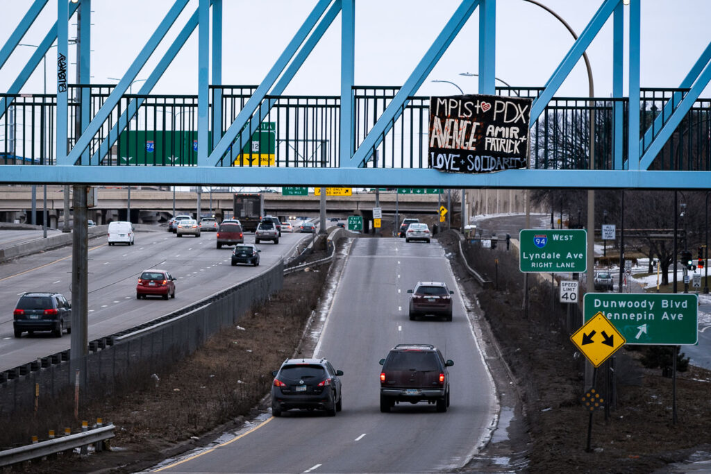 A banner hangs from the Irene Hixon Whitney Bridge during rush hour outside the Lowry Hill Tunnel. The sign reads MPLS PDX AVENGE LOVE SOLIDARITY and was dropped a day after protesters were fired upon in Portland where they were marching for Amir Locke.