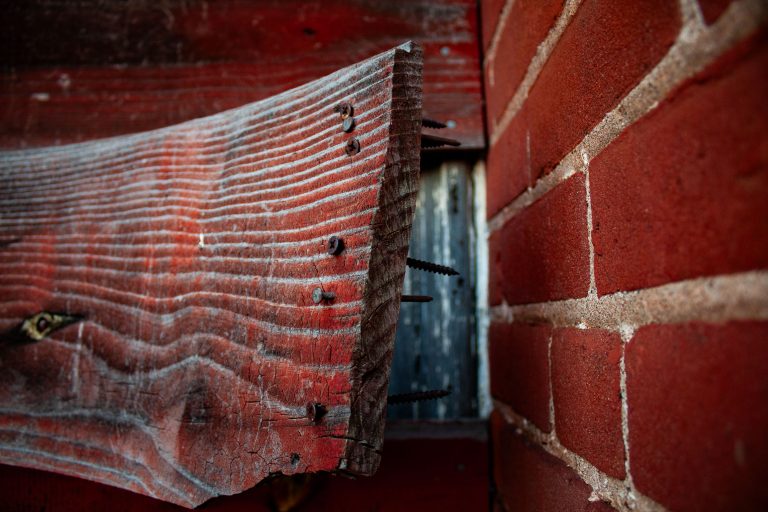 Exposed Nails in Weathered Wood on Brick Wall 1 Weathered red wood with protruding nails is attached to a brick wall on an abandoned building.