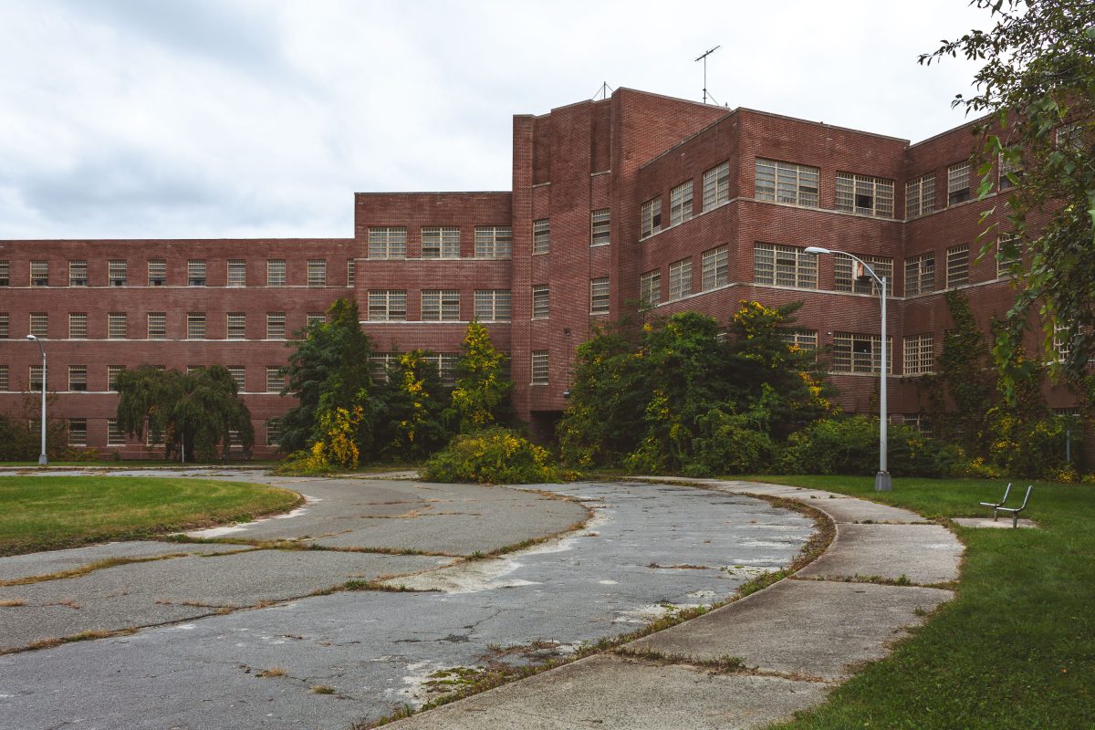 Abandoned brick institutional building with barred windows surrounded by overgrown vegetation and deteriorating pavement at Kings