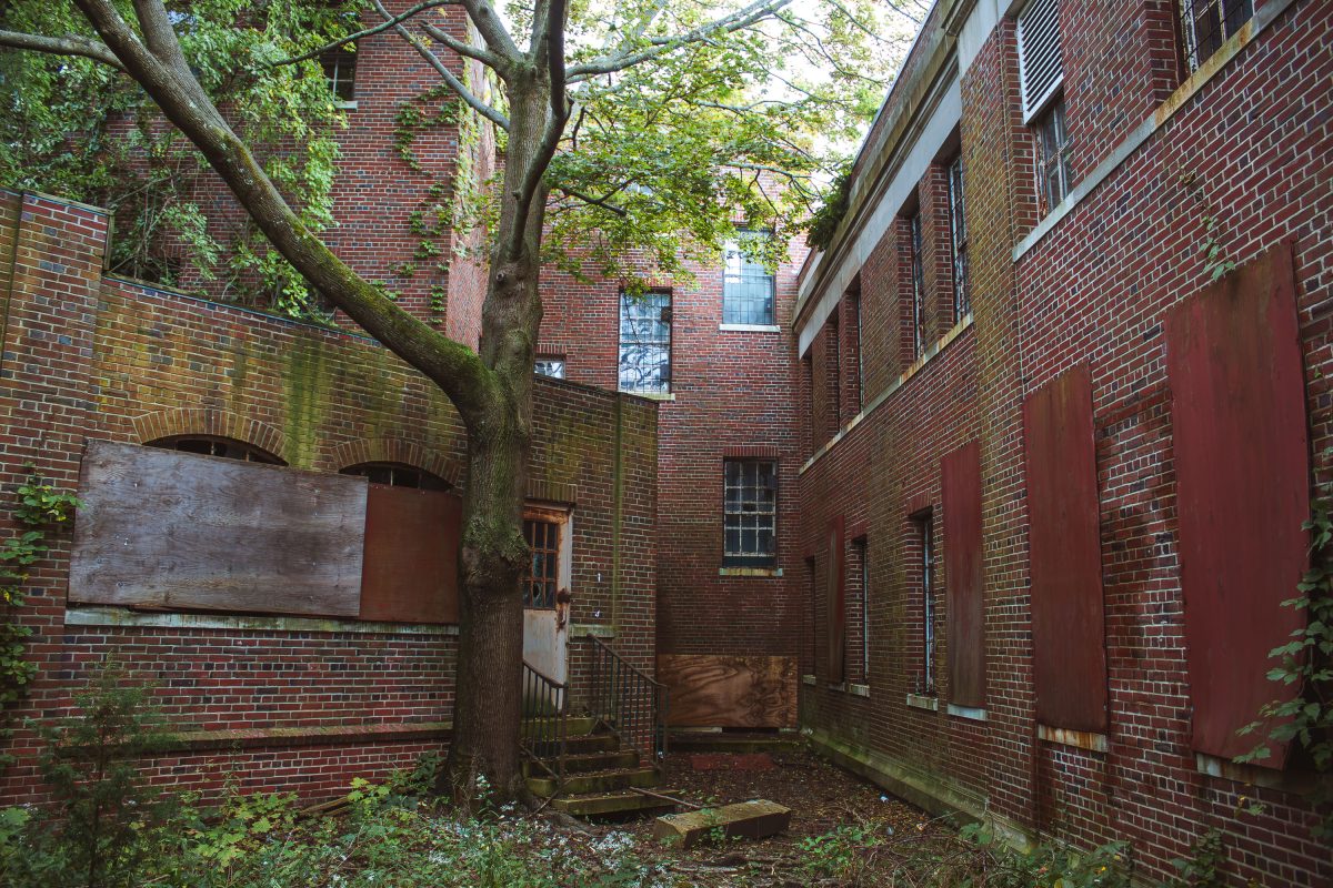Overgrown courtyard between red brick buildings with barred windows at Kings Park Psychiatric Center, New York.