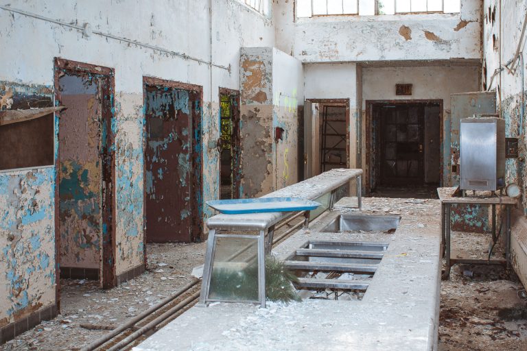 Communal Sink Trough and Ward Corridor, Kings Park Psychiatric Center 3 Abandoned institutional hallway with rusted metal examination table and deteriorating blue-painted walls at Kings Park
