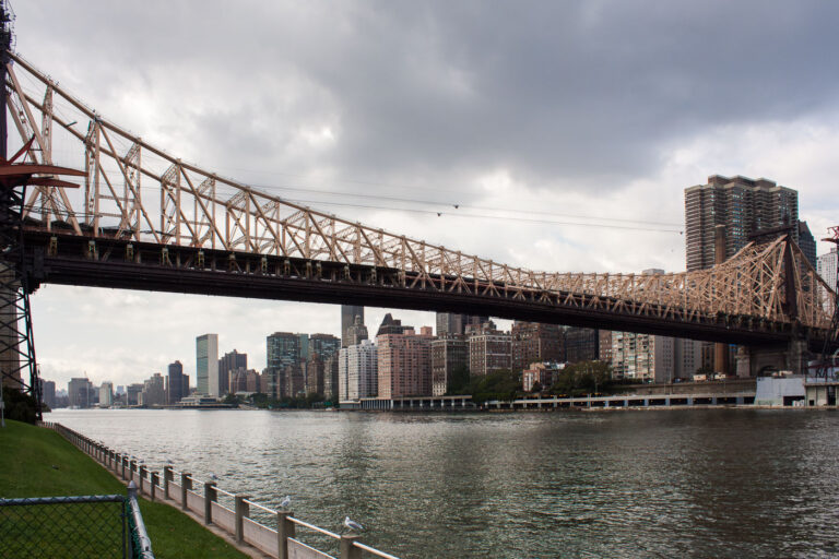 Ed Koch Queensboro Bridge over the East River 4 The Ed Koch Queensboro Bridge, spanning the East River between Manhattan and Queens, is captured here under an overcast sky from Roosevelt Island. Completed in 1909, this double-deck cantilever bridge remains one of New York City’s most significant engineering landmarks, carrying both vehicular and pedestrian traffic while offering a vital connection between Midtown Manhattan and Long Island City. Above the roadway, the Roosevelt Island Tram cables can be seen tracing parallel to the structure — a modern counterpart to this early 20th-century feat of steelwork. The muted light of the cloudy day accentuates the warm tones of the bridge’s framework against the cool gray skyline of Manhattan.