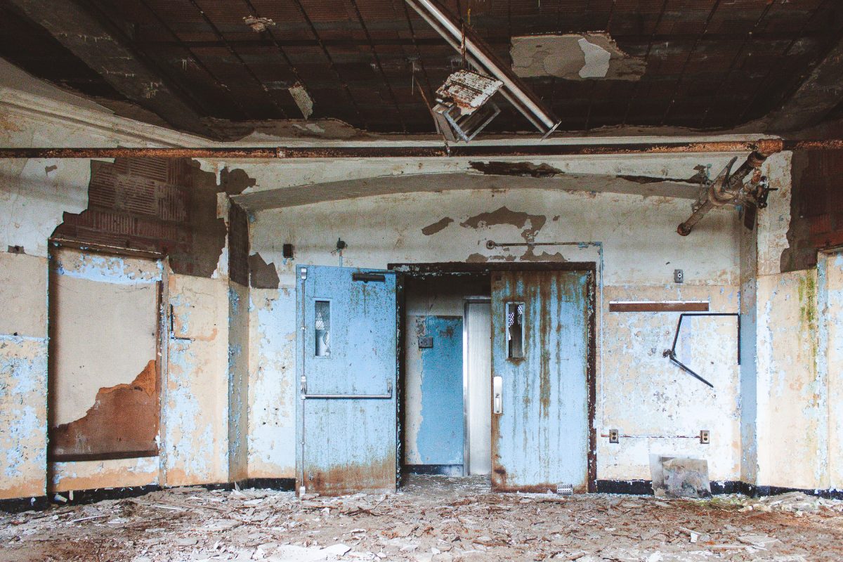 Abandoned institutional hallway with peeling blue paint, rusted doors, and collapsed ceiling at Kings Park Psychiatric Center.