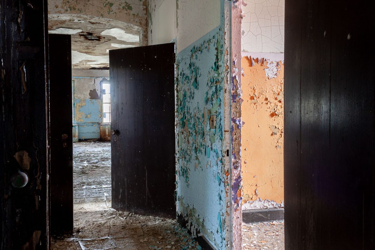 Deteriorated hallway with open dark doors and peeling blue, orange, and white paint at Kings Park Psychiatric Center.