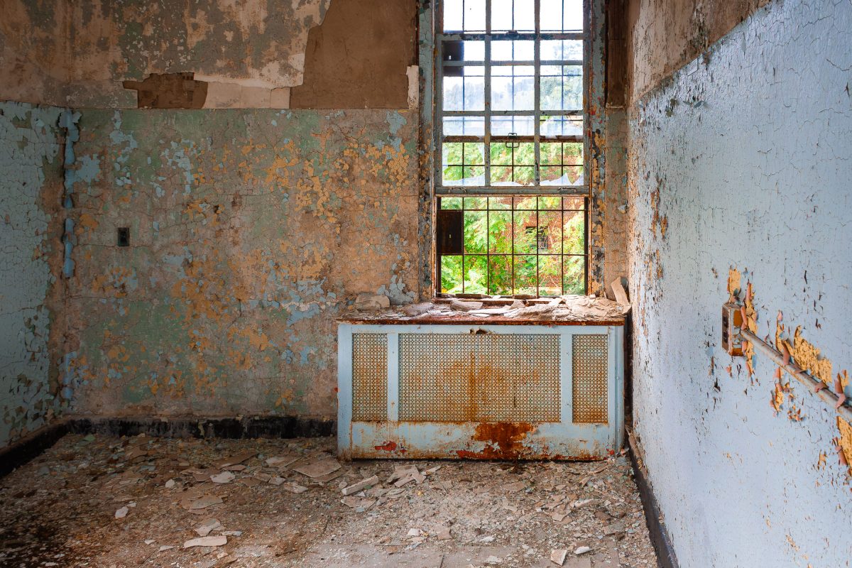 Abandoned institutional room with deteriorating walls, rusted radiator, and barred window at Kings Park Psychiatric Center.