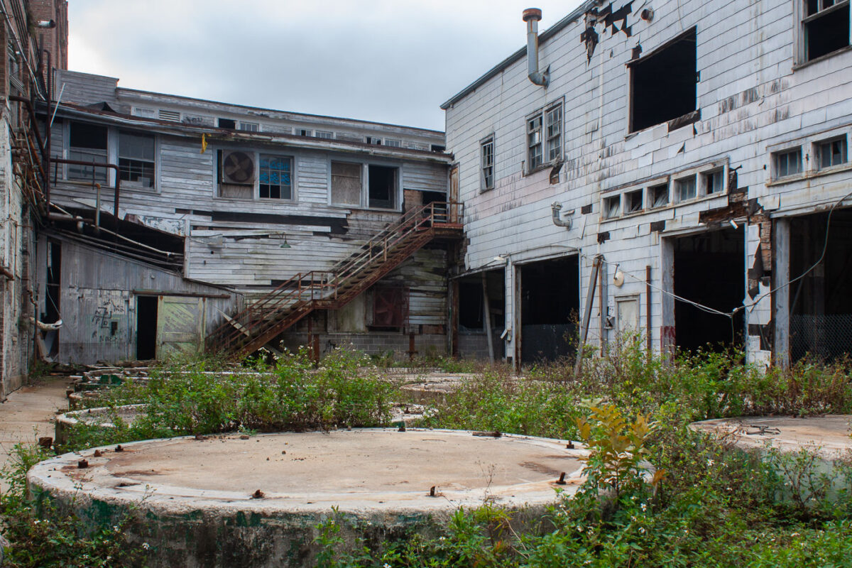 Abandoned Dixie Brewery buildings in New Orleans show decay with weathered siding, broken windows, and overgrown grounds. Graffiti is visible on structures.