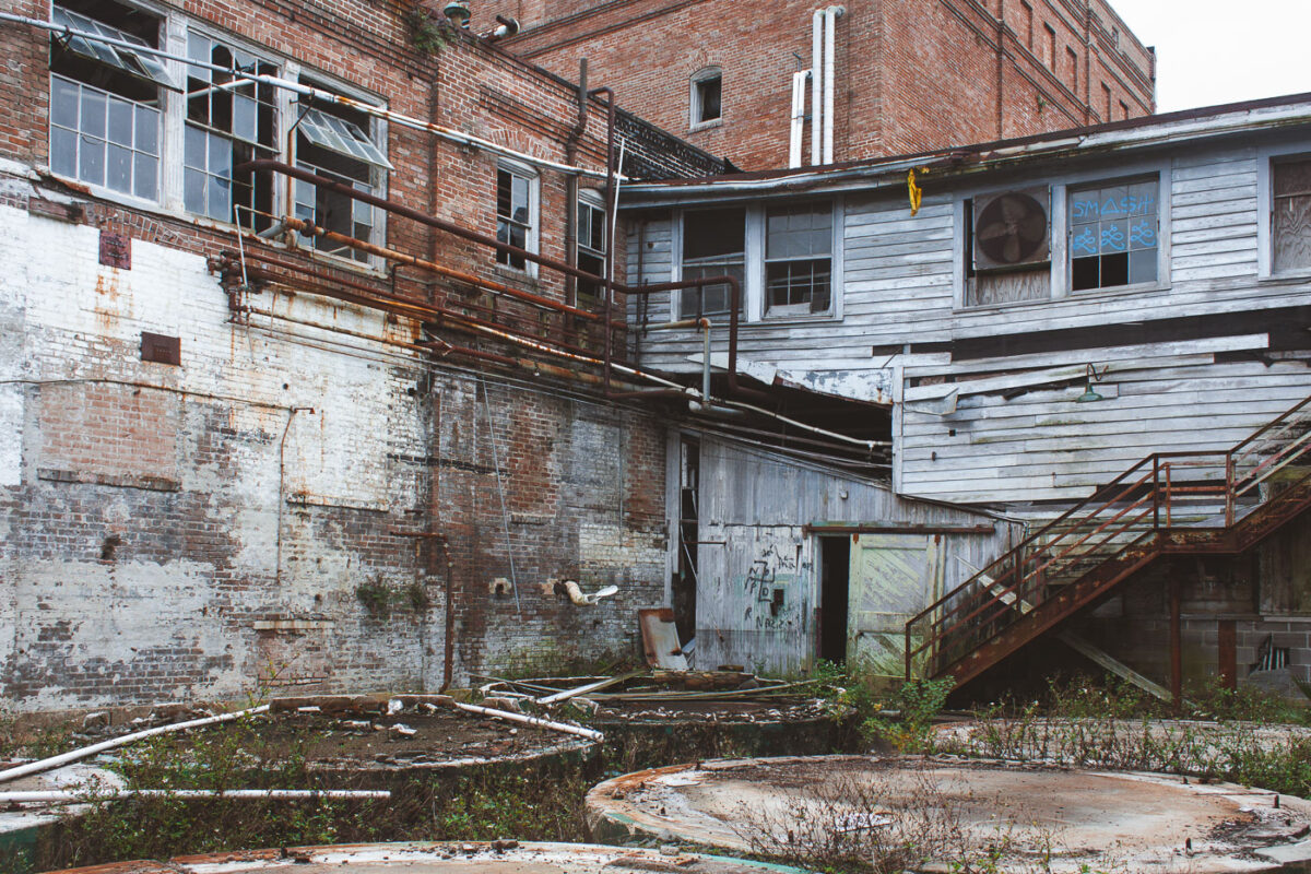 The abandoned Dixie Brewery in New Orleans, a historic industrial landmark, shows signs of decay with weathered brick, peeling paint, broken windows, and overgrown vegetation.