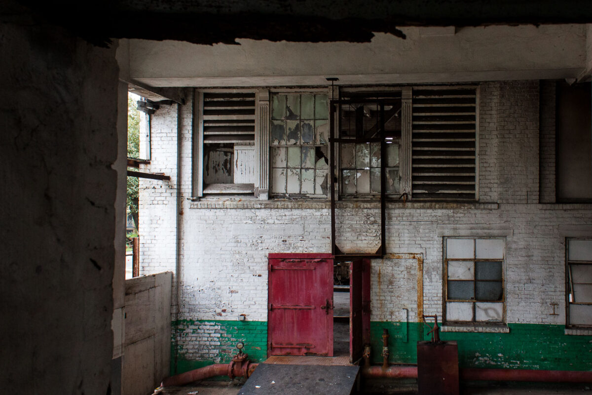 Interior of the abandoned Dixie Brewery in New Orleans, showing peeling white brick walls, broken windows, industrial piping, and a red door.
