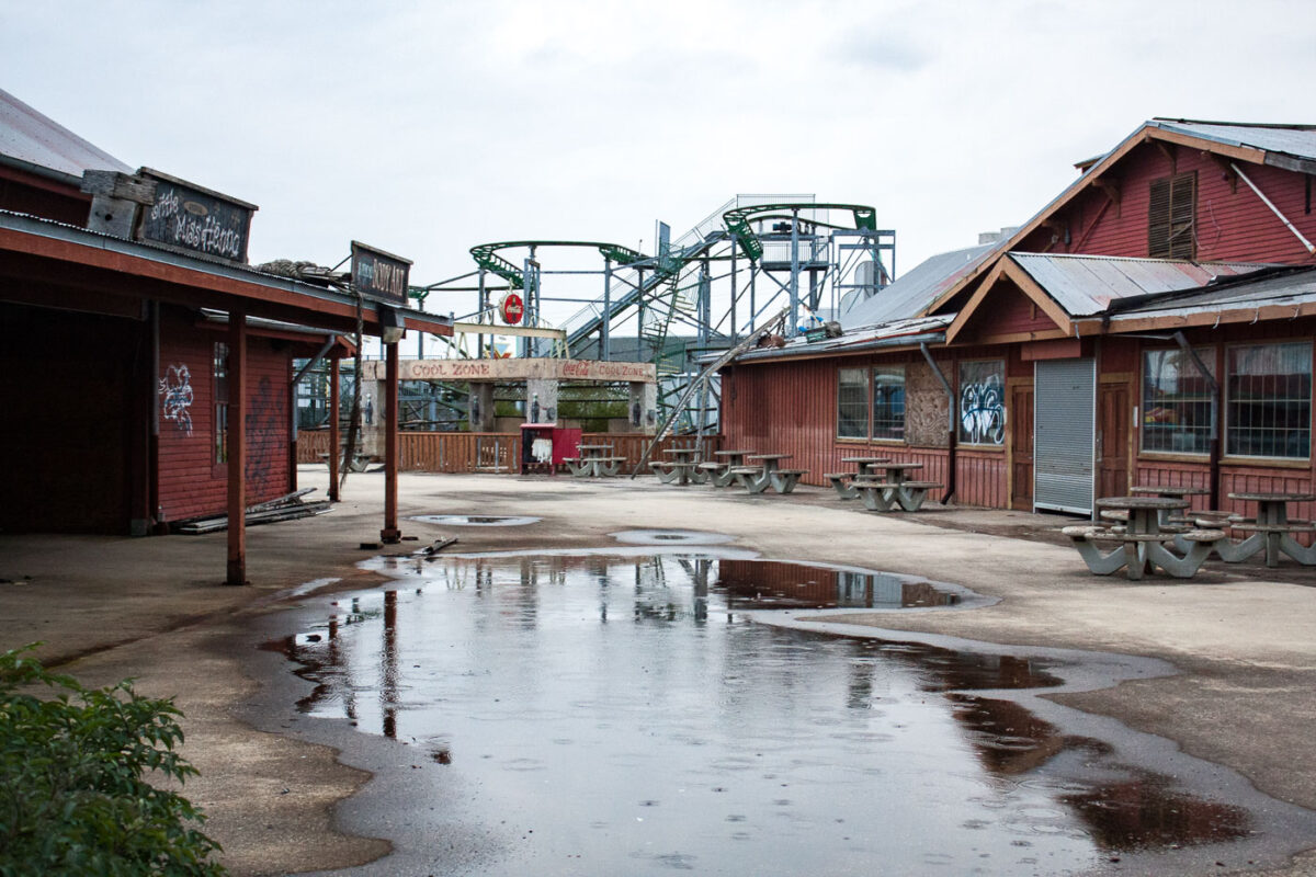 The abandoned Six Flags New Orleans amusement park, damaged by Hurricane Katrina, shows decay with graffiti and puddles. A roller coaster is visible in the background.
