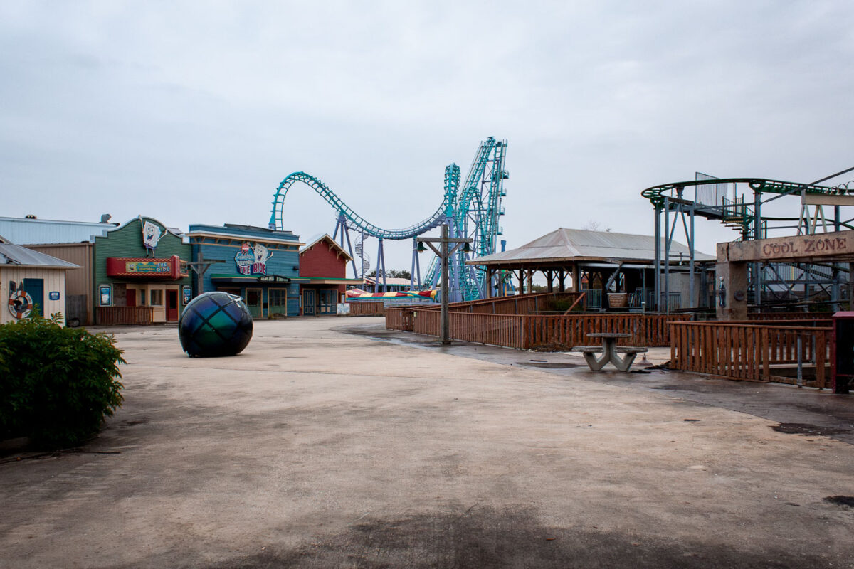 The abandoned Six Flags New Orleans amusement park, damaged by Hurricane Katrina, shows overgrown vegetation and a prominent roller coaster. A "SpongeBob SquarePants The Ride" building is visible.