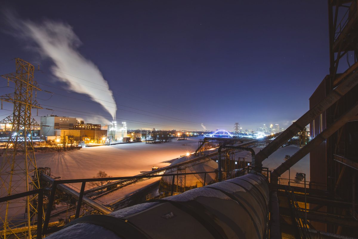 Upper Harbor Terminal barge facility on the frozen Mississippi River in Minneapolis at dusk, with downtown skyline and Stone Arch Bridge visible in background.