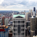 Prominent in the foreground is 301 South Wacker Drive, completed in 1989 in a postmodern style, identifiable by its green pyramidal roof and vertical glass grid patterns that reflected late-20th-century corporate architectural design in Chicago. Just to the right are the Marina City towers, completed in 1967 by architect Bertrand Goldberg as one of the earliest large-scale mixed-use residential developments intended to draw residents back into the urban core during a period of downtown population decline.

Farther north, the tall dark building with twin antenna masts is the John Hancock Center, completed in 1969 using innovative bundled-tube engineering developed in Chicago, a system that allowed greater height and wind resistance while reducing material use. Beyond it, the Lake Michigan shoreline reveals Chicago’s growth toward the lakefront, where residential high-rises expanded throughout the 1970s and 1980s to serve tourism, business housing, and dense urban living. The view illustrates the evolution of Chicago’s skyline as a center of architectural experimentation and urban redevelopment.