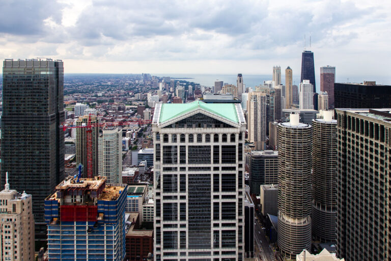 Chicago Architecture and Lake Michigan 1 Prominent in the foreground is 301 South Wacker Drive, completed in 1989 in a postmodern style, identifiable by its green pyramidal roof and vertical glass grid patterns that reflected late-20th-century corporate architectural design in Chicago. Just to the right are the Marina City towers, completed in 1967 by architect Bertrand Goldberg as one of the earliest large-scale mixed-use residential developments intended to draw residents back into the urban core during a period of downtown population decline.Farther north, the tall dark building with twin antenna masts is the John Hancock Center, completed in 1969 using innovative bundled-tube engineering developed in Chicago, a system that allowed greater height and wind resistance while reducing material use. Beyond it, the Lake Michigan shoreline reveals Chicago’s growth toward the lakefront, where residential high-rises expanded throughout the 1970s and 1980s to serve tourism, business housing, and dense urban living. The view illustrates the evolution of Chicago’s skyline as a center of architectural experimentation and urban redevelopment.
