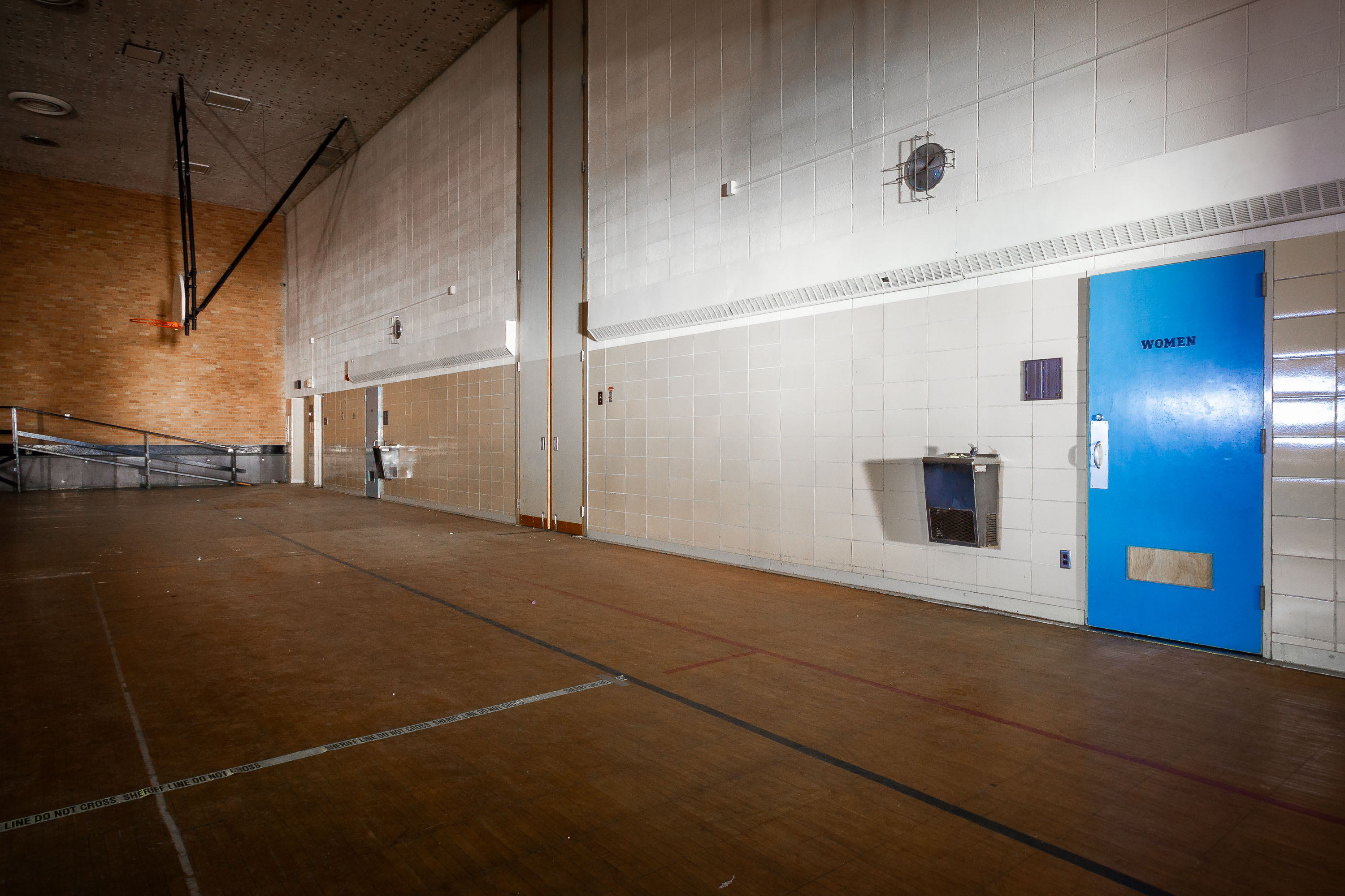Abandoned institutional hallway with blue women's restroom door and white tile walls at Eagle Island Psychiatric Hospital.