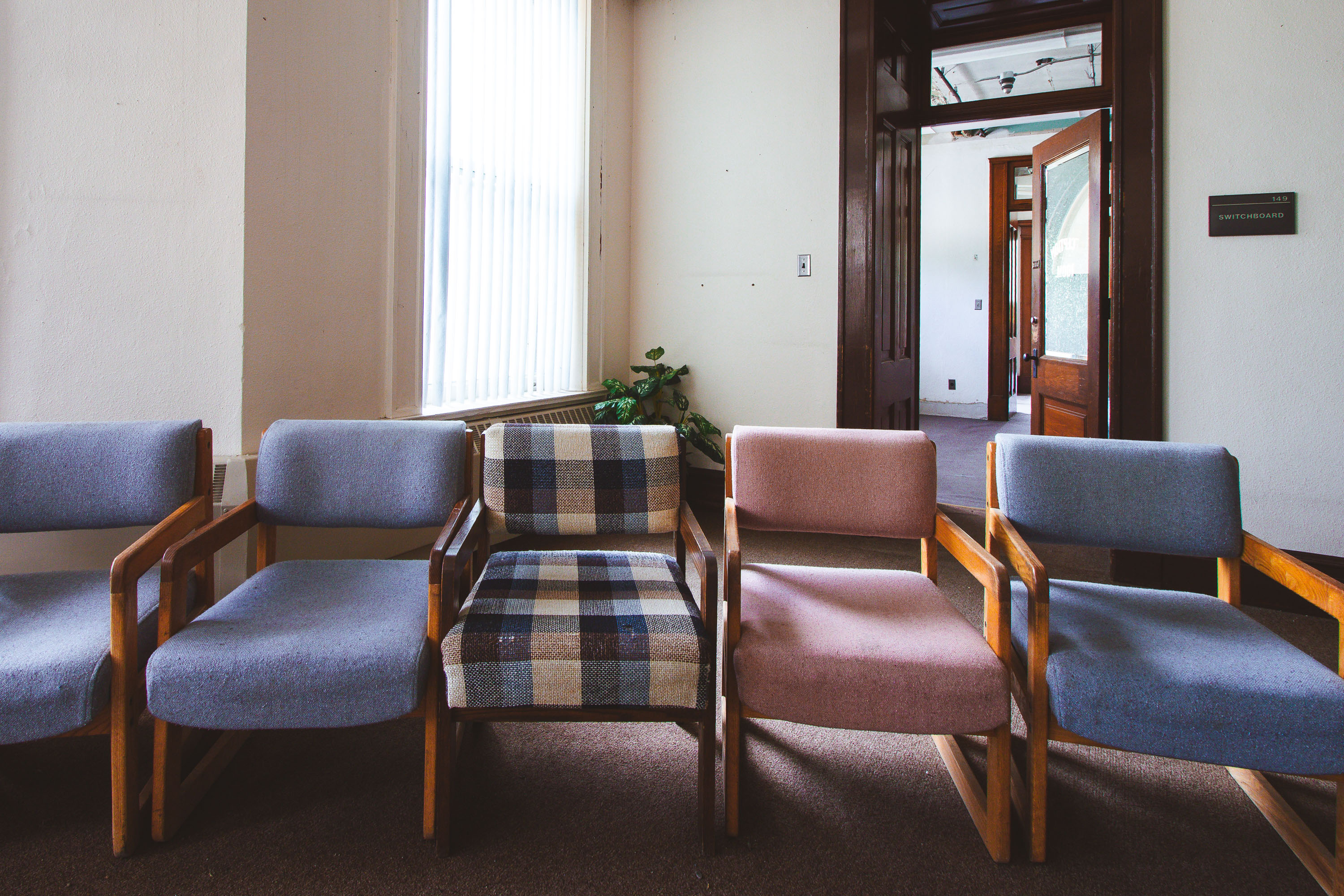 Empty waiting room with five wooden-framed chairs in blue and plaid upholstery inside a institutional building.