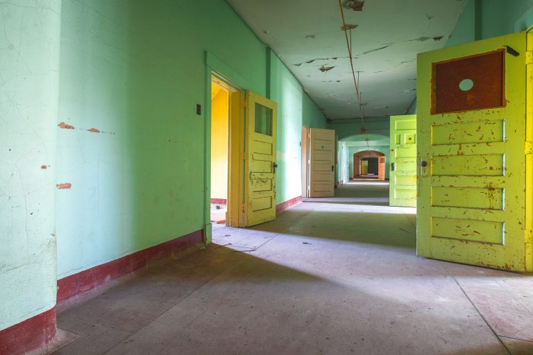 Ward corridor with painted doors, Fergus Falls Kirkbride Building 4 Abandoned institutional corridor with green walls, yellow doors, and rust-stained surfaces at Eagle Island Psychiatric Hospital.