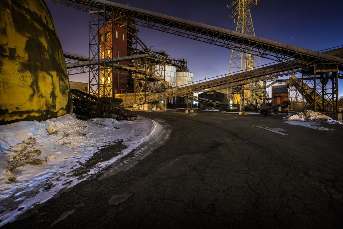 Industrial barge terminal with illuminated metal structures, conveyor systems, and equipment along the Mississippi River in north Minneapolis at night.