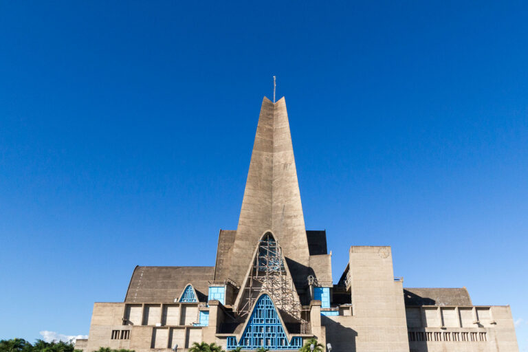 Basilica of Our Lady of Altagracia, Higüey 1 The Basílica Catedral Nuestra Señora de la Altagracia in Higüey, Dominican Republic, is one of the most significant Catholic pilgrimage destinations in the Caribbean. Completed in 1971, the church was designed by French architects André-Jacques Dunoyer de Segonzac and Pierre Dupré, who created a unique modernist interpretation of a basilica with towering concrete arches and geometric symmetry meant to evoke hands raised in prayer. The blue metal-framed windows contrast against the stark reinforced concrete, emphasizing both spiritual and structural purpose. The basilica was commissioned to honor the Virgin of Altagracia, protector of the Dominican people, and it officially received the status of Minor Basilica from Pope Paul VI. Known for its 80-meter bell tower and monumental entry arch, it attracts thousands of worshippers every January 21 during the Feast of Altagracia.
