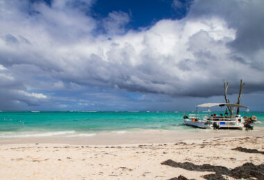 Along the eastern coast of Punta Cana in the Dominican Republic, a coastal tour boat rests against the pale sandy shore as shifting weather approaches over the Caribbean Sea. The turquoise shallows—characteristic of this stretch of the Atlantic coast lined by coral reefs—contrast sharply with a dense bank of darkening storm clouds moving inland. Beach excursions and snorkeling trips commonly depart from this area, where the water remains shallow and calm before the reef break. The photograph captures the transitional weather common to the region’s tropical maritime climate, highlighting the tension between the inviting clarity of the shoreline and the dramatic sky signaling an imminent rainfront.