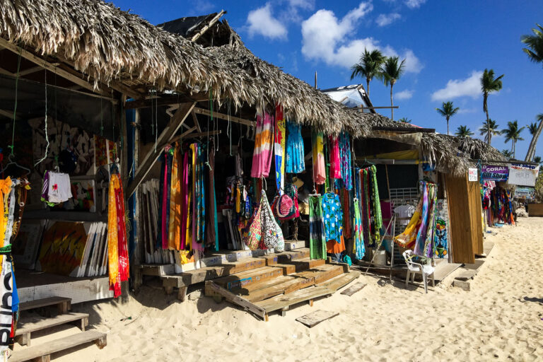 Beach vendors near Iberostar Selection Hacienda Dominicus 2 Colorful sarongs, paintings, handmade bags, and souvenirs hang from rustic palm-thatched stalls built directly over the white sand near Iberostar Selection Hacienda Dominicus in southeastern Dominican Republic. These informal beach vendors are part of the longstanding tourism-driven micro-economy that developed along the shoreline between Bayahibe and Dominicus Americanus, where local artisans sell crafts to resort visitors. The improvised wooden structures—some with painted steps and corrugated roofing—reflect the region’s blend of tourism and local entrepreneurship, an essential livelihood for many residents in La Altagracia Province.