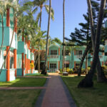Hotel El Cortecito Courtyard Lodgings, Punta Cana 3 Colorful courtyard lodging buildings at Hotel El Cortecito in Punta Cana, Dominican Republic. Situated in the El Cortecito beach district—one of the earliest tourism hubs in the area—this hotel reflects the architectural style common in locally-owned coastal accommodations before the rise of large all-inclusive resorts. The turquoise and terracotta exterior, white balustrades, and open garden walkways offer a Caribbean vernacular blend intended for natural ventilation and communal outdoor space. El Cortecito developed through the 1980s and 1990s as a fishing village turned tourism enclave, with modest inns like this one often built and expanded in phases by local families, contrasting with the master-planned resort complexes that later defined Punta Cana’s international tourism growth.
