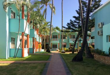 Colorful courtyard lodging buildings at Hotel El Cortecito in Punta Cana, Dominican Republic. Situated in the El Cortecito beach district—one of the earliest tourism hubs in the area—this hotel reflects the architectural style common in locally-owned coastal accommodations before the rise of large all-inclusive resorts. The turquoise and terracotta exterior, white balustrades, and open garden walkways offer a Caribbean vernacular blend intended for natural ventilation and communal outdoor space. El Cortecito developed through the 1980s and 1990s as a fishing village turned tourism enclave, with modest inns like this one often built and expanded in phases by local families, contrasting with the master-planned resort complexes that later defined Punta Cana’s international tourism growth.