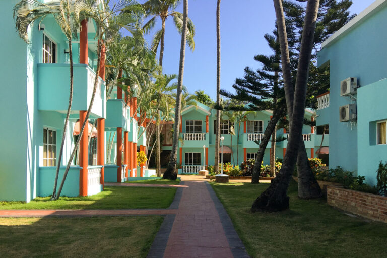 Hotel El Cortecito Courtyard Lodgings, Punta Cana 3 Colorful courtyard lodging buildings at Hotel El Cortecito in Punta Cana, Dominican Republic. Situated in the El Cortecito beach district—one of the earliest tourism hubs in the area—this hotel reflects the architectural style common in locally-owned coastal accommodations before the rise of large all-inclusive resorts. The turquoise and terracotta exterior, white balustrades, and open garden walkways offer a Caribbean vernacular blend intended for natural ventilation and communal outdoor space. El Cortecito developed through the 1980s and 1990s as a fishing village turned tourism enclave, with modest inns like this one often built and expanded in phases by local families, contrasting with the master-planned resort complexes that later defined Punta Cana’s international tourism growth.