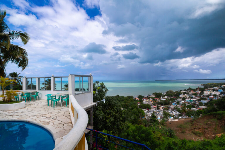 Hotel La Loma Overlooking Miches and Laguna Redonda 1 Perched high on one of the hills surrounding the coastal town of Miches in eastern Dominican Republic, Hotel La Loma offers a panoramic view of the shoreline, the town below, and the wide expanse of Laguna Redonda stretching toward the horizon. The hotel’s elevated terrace and pool area, with simple seating and curved white railings, reflects the region’s small-scale tourism development before large resort expansion began nearby in the late 2010s. From this vantage point, you can see Miches’ compact urban fabric, local cemetery, and fishing boats that support the town’s traditional economy—contrasting with growing interest in ecotourism and coastal resort projects along the north shore. The turquoise water of the protected lagoon and the dramatic storm clouds illustrate the area’s tropical climate, shaped by trade winds and sudden rainfall typical of the Dominican northeast.