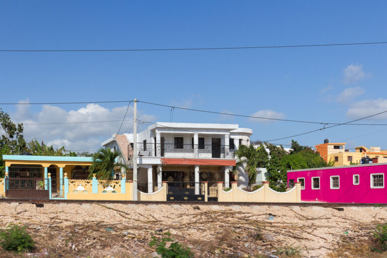 La Romana, Dominican Republic: Colorful Architecture by Railroad 1 Residential buildings in La Romana, Dominican Republic, are situated adjacent to a railway line. The architecture displays a variety of styles and vibrant colors, with the prominent white structure featuring classical columns and balconies. The presence of the railroad suggests a historical connection to transportation and industry in the region, likely for the movement of goods or people. Today, these structures serve as homes, reflecting the contemporary urban landscape of La Romana.