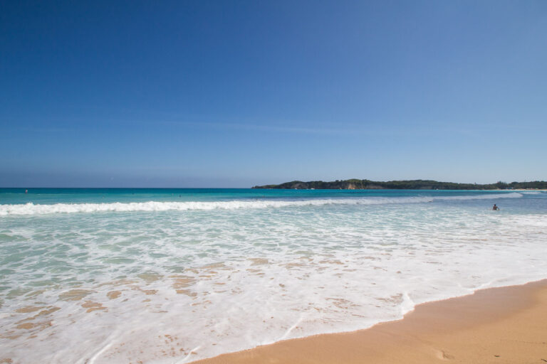 Shoreline Calm at Macao Beach 1 Along the northeastern coast of the Dominican Republic, Macao Beach is one of the rare stretches of undeveloped shoreline in the Punta Cana region. Unlike most resort-dominated beaches of the eastern coast, Macao has remained publicly accessible, attracting both local residents and visitors for its surfing conditions, expansive sands, and panoramic views of the Atlantic. The gently rolling waves seen here are typical of the beach’s surfable breaks, shaped by offshore currents and an open coastline without coral barriers.
In the distance, the low, wooded hills form the edge of the Punta Cana–Miches coastal corridor, an area where farmland, mangroves, and small fishing communities meet tourism expansion. Known historically as a landing spot for local fishermen, Macao has also been a training and gathering place for Dominican surfing youth. Today it marks a contrast between traditional coastal use and the rapid rise of resort development across the eastern Dominican shoreline.