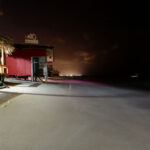 structure, with a palm-thatch roof and simple seating, reflects the informal beachfront culture of the region, where many bars operate just steps from the water. Captured at night with a long exposure, the Caribbean Sea appears smooth and misty as waves gently wash beneath the structure. In the distance, faint shoreline lights show the resort-lined coast of Punta Cana. The photo highlights the fragility of beachfront structures, many of which face increasing erosion and tidal encroachment due to coastal development and rising sea levels.