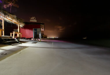 structure, with a palm-thatch roof and simple seating, reflects the informal beachfront culture of the region, where many bars operate just steps from the water. Captured at night with a long exposure, the Caribbean Sea appears smooth and misty as waves gently wash beneath the structure. In the distance, faint shoreline lights show the resort-lined coast of Punta Cana. The photo highlights the fragility of beachfront structures, many of which face increasing erosion and tidal encroachment due to coastal development and rising sea levels.