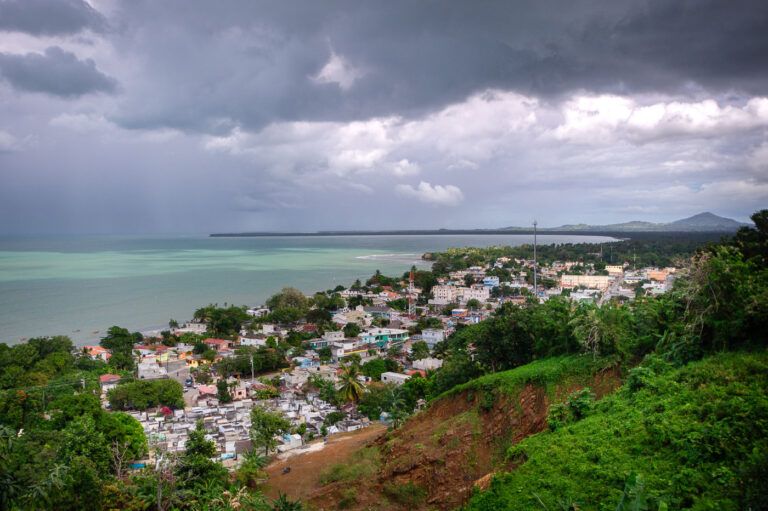 Stormy Coastal View over Miches, Dominican Republic 2 Elevated above the small town of Miches on the northeastern coast of the Dominican Republic, this vantage point offers a sweeping view of the Caribbean shoreline and the lush countryside of El Seibo Province. The town’s clustered homes, cemetery, and local roads sit beneath dramatic rain clouds moving in from the sea, while the pale turquoise water hints at the nearby shallow coastal shelf. Miches has historically relied on fishing and agriculture, but over the past decade it has become a gateway to expanding tourism projects along the coastal corridor toward Punta Cana and Samaná. From this hillside, the contrast between traditional local development and the region’s growing transformation is clearly visible against the backdrop of changing skies and the mountainous horizon.