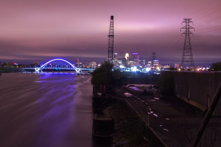 Lowry Avenue Bridge Seen From Upper Harbor Terminal at Night 2 The Stone Arch Bridge illuminates blue over the Mississippi River at dusk, with Minneapolis downtown skyline and power transmission towers beyond.