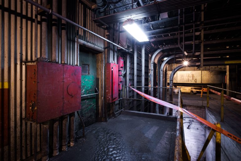 Industrial corridor inside ArcelorMittal steel plant with red metal doors, metal railings, and overhead lighting.
