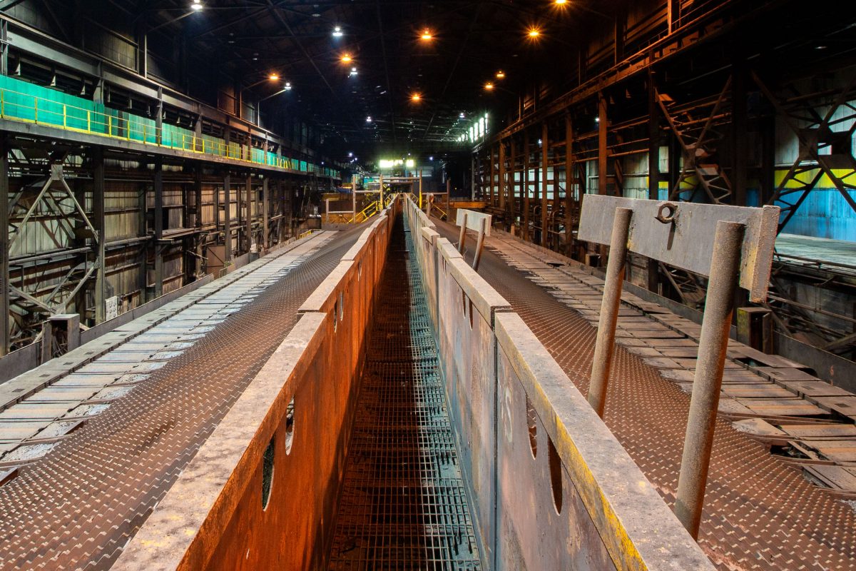Nighttime view of an industrial steel mill facility with an orange operator's walkway, metal railings, and overhead industrial infrastructure.