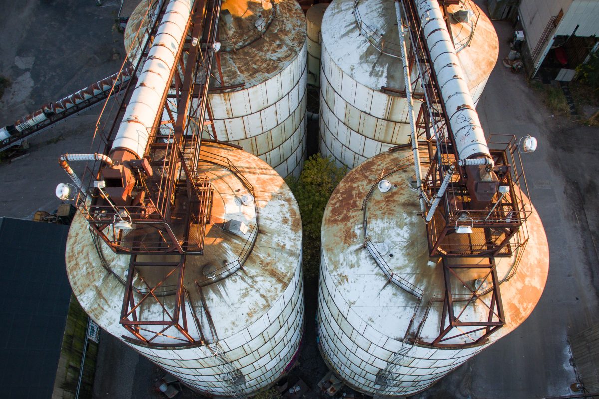 Aerial view of two large cylindrical storage tanks with rusted tops and metal loading structures at Upper Harbor Terminal on the Mississippi River in Minneapolis.