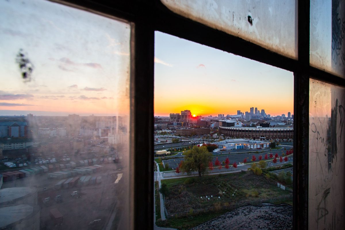 Minneapolis skyline at sunset viewed through an abandoned industrial building's window frame.