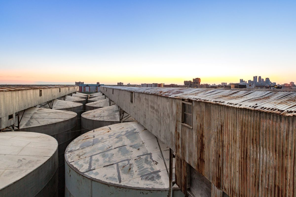 Abandoned Electric Steel grain elevator rooftop in Minneapolis with large storage tanks and downtown skyline at sunset.