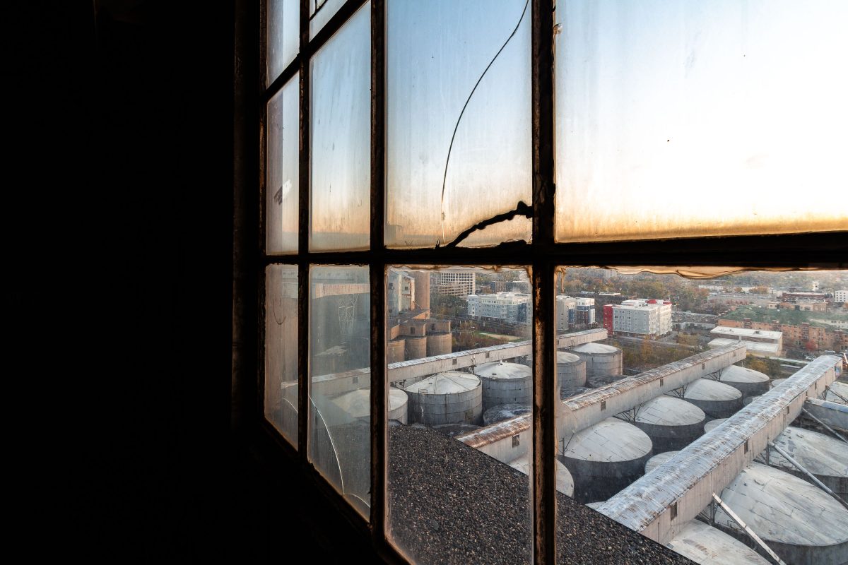 View from inside an abandoned Minneapolis grain elevator looking out at storage tanks and the city skyline at sunset.
