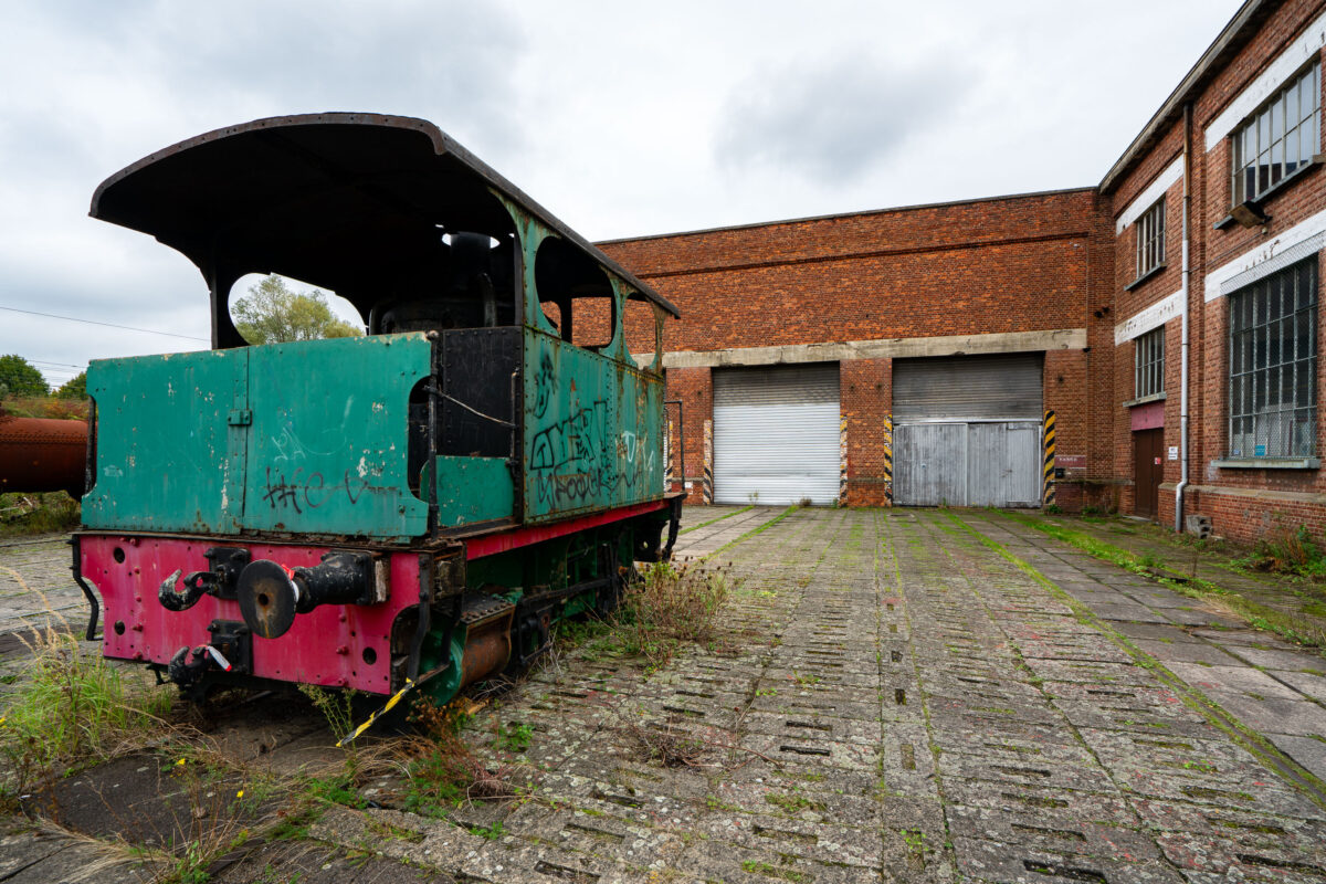 Abandoned locomotive at an industrial site in Belgium, with a brick building in the background.