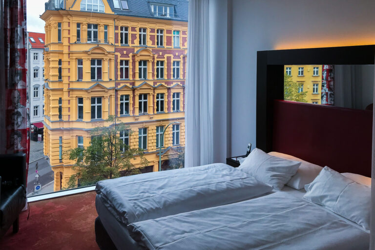 A modern hotel room in Berlin’s Mitte district looks out through a floor‑to‑ceiling window onto a restored 19th‑century residential block across the street. The room features two neatly made single beds with white linens, a dark-framed headboard with soft backlighting, a bedside table with a phone, and sheer curtains patterned with portrait graphics. Outside, the opposite building displays yellow stucco and red-brick detailing typical of Gründerzeit architecture, with tall windows, cornices, and balconies above a tree‑lined sidewalk. The scene is consistent with accommodations such as the Arcotel Velvet Berlin, located along a corridor of cafés, galleries, and tram routes that connect the Mitte neighborhood with nearby cultural sites. The image reflects the mix of contemporary hospitality design and historic streetscapes that characterize central Berlin decades after reunification.