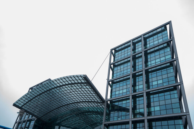 Berlin Hauptbahnhof Berlin Central Station 4 The glass-and-steel facade of Berlin Hauptbahnhof rises under an overcast sky in Berlin, Germany. The image shows the arched glass roof canopy anchored by steel cables and a rectilinear tower with blue‑tinted window grids, part of the station’s multi-level entrance complex. Located in the Mitte district near the government quarter and the River Spree, the station opened in 2006 on the former site of Lehrter Bahnhof as a flagship post‑reunification infrastructure project. Today it is a major hub for Deutsche Bahn’s ICE and regional services, with S‑Bahn and the U5 metro connection below, linking north–south and east–west routes across the capital and beyond.