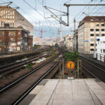 Berlin Friedrichstraße train station S Bahn Tracks 2 View east from Berlin Friedrichstraße station showing the elevated S-Bahn tracks leading toward Alexanderplatz. Overhead wires and supporting steel structures frame the route, with graffiti-covered industrial buildings on the left and mid-century offices on the right. The Berlin TV Tower (Fernsehturm) rises in the distance, a landmark visible across the city skyline.