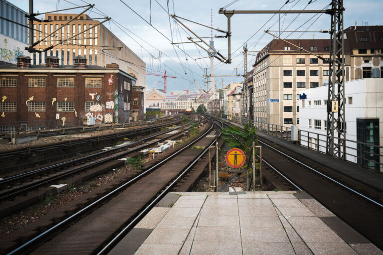 Berlin Friedrichstraße train station S Bahn Tracks 3 View east from Berlin Friedrichstraße station showing the elevated S-Bahn tracks leading toward Alexanderplatz. Overhead wires and supporting steel structures frame the route, with graffiti-covered industrial buildings on the left and mid-century offices on the right. The Berlin TV Tower (Fernsehturm) rises in the distance, a landmark visible across the city skyline.