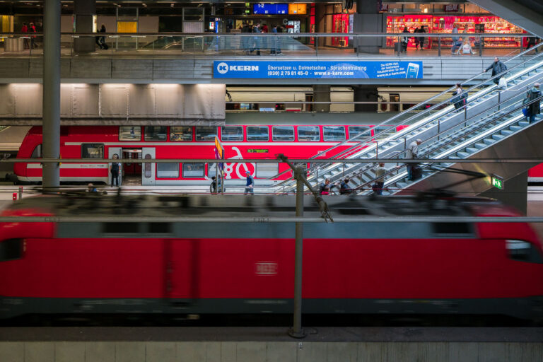 Berlin Hauptbahnhof - Berlin Central Station 1 Passengers move across multiple levels inside Berlin Hauptbahnhof, Germany’s central rail hub in the capital. A red double-decker Regionalbahn train stands at a middle platform with doors open, marked by a large bicycle symbol on one carriage. A red Deutsche Bahn locomotive blurs past on a lower track, while travelers ride escalators that cut diagonally through the scene. Above, a retail concourse includes a Relay shop and a blue banner advertising KERN language services, with platform indicators for tracks 1–2 visible. Opened in 2006 on the site of the former Lehrter Bahnhof, Berlin Central Station is a major interchange for regional, long‑distance, and S‑Bahn services in Germany.