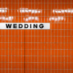 A white station sign reading “Wedding” in black block letters is mounted on an orange, small-rectangle tiled wall inside Wedding station in Berlin, Germany. The station serves the Wedding neighborhood in the Mitte district and functions as an interchange between the U6 U‑Bahn line and the Berlin Ringbahn S‑Bahn. The stark typography and modular tiles reflect the practical design found across much of Berlin’s postwar transit architecture. Operated by BVG for the U‑Bahn and S‑Bahn Berlin GmbH for suburban rail, Wedding is a local hub for commuters moving between the north of the city and central Berlin. No passengers are visible in the frame, emphasizing the graphic clarity of the station’s wayfinding.