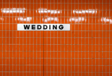 A white station sign reading “Wedding” in black block letters is mounted on an orange, small-rectangle tiled wall inside Wedding station in Berlin, Germany. The station serves the Wedding neighborhood in the Mitte district and functions as an interchange between the U6 U‑Bahn line and the Berlin Ringbahn S‑Bahn. The stark typography and modular tiles reflect the practical design found across much of Berlin’s postwar transit architecture. Operated by BVG for the U‑Bahn and S‑Bahn Berlin GmbH for suburban rail, Wedding is a local hub for commuters moving between the north of the city and central Berlin. No passengers are visible in the frame, emphasizing the graphic clarity of the station’s wayfinding.